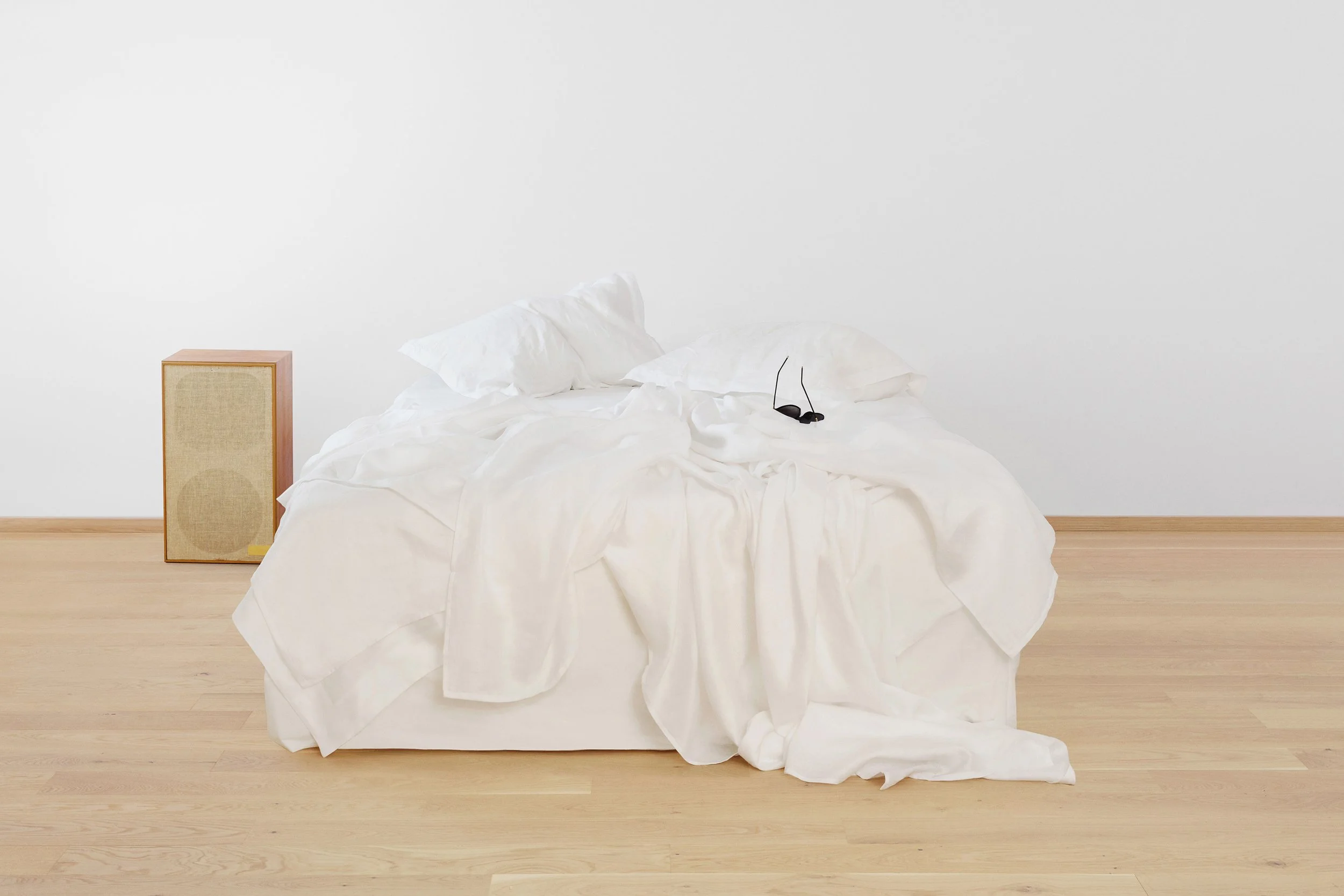 A bed with a crumpled white sheet and two pillows, black sunglasses resting on the sheet, beige speaker on the left, and plain white wall in the background.
