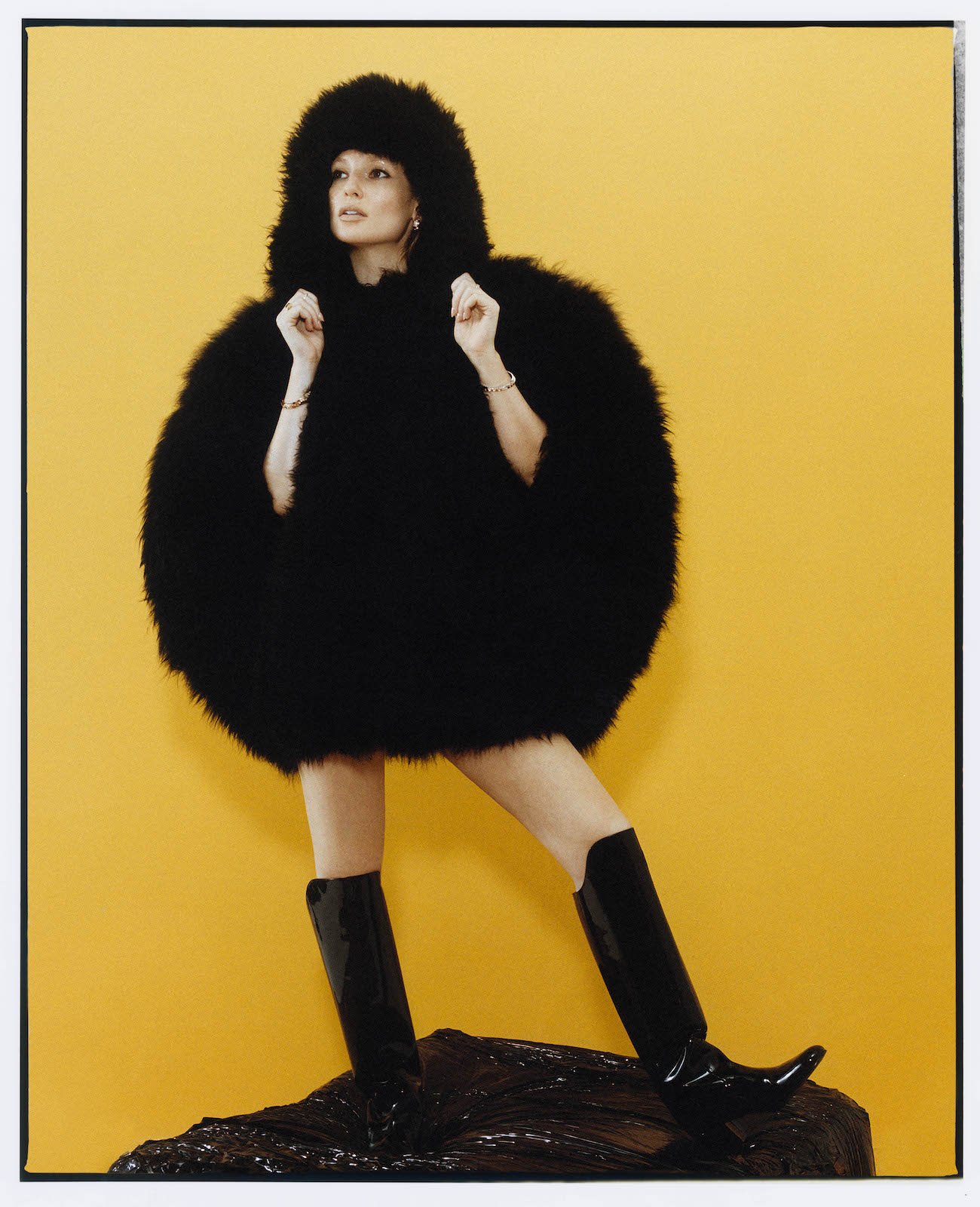 Fashion model wearing a large black fur coat with hood, black boots, posing against a yellow background, standing on a black textured surface.