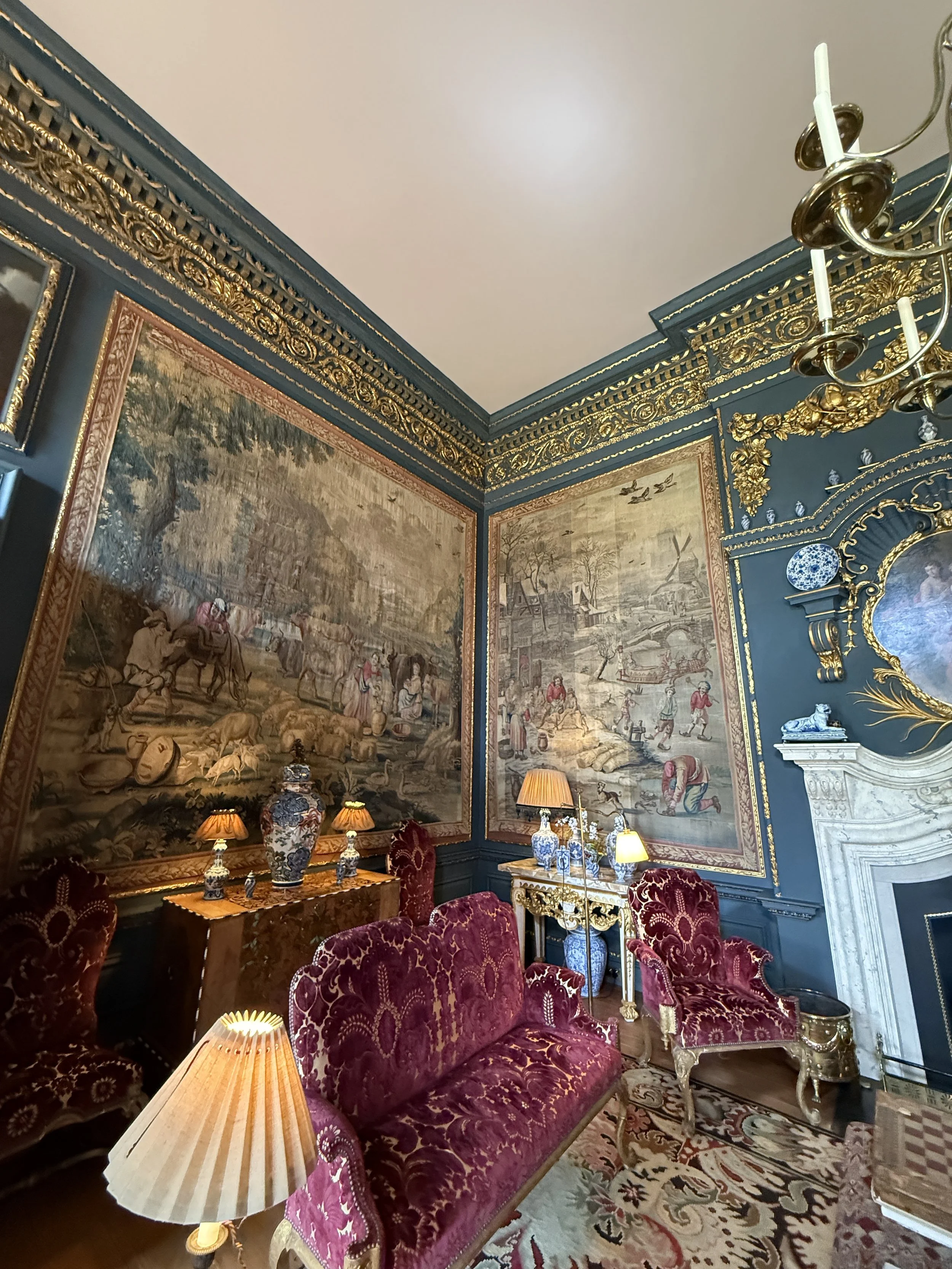 A richly decorated sitting room with dark blue walls and ornate gold trim. Large tapestries depicting historical scenes hang on the walls. There is a velvet upholstered sofa in a deep red hue and matching chairs, with a gold frame. Several table lamps are on a wooden table and a side table, illuminating the space. A white marble fireplace with intricate moldings is on the right wall, topped with a small sculpture and a blue and white decorative plate. The ceiling is painted white, with a gold chandelier hanging from it. The room has a patterned area rug on the wooden floor.