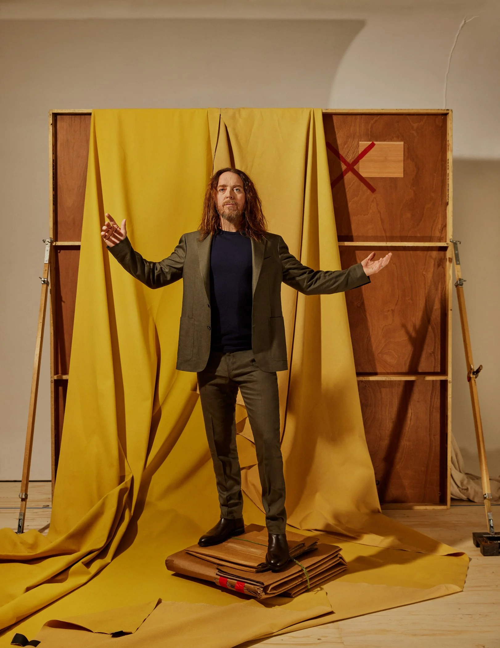 Comedian Tim Minchin standing with his arms outstretched in front of a backdrop of yellow curtains, with a wooden structure and fabric surrounding him. For the Guardian newspaper.