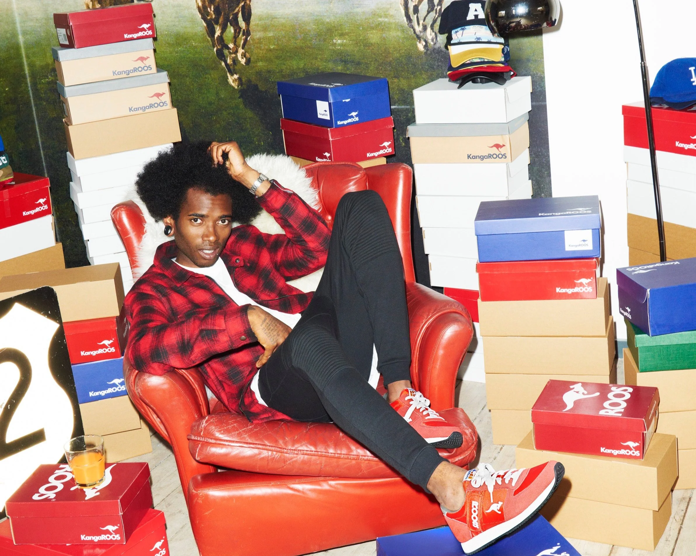 A young man reclining on a red leather couch surrounded by stacks of shoe boxes in various colors, with a background of a mural of giraffes and grassy plains.
