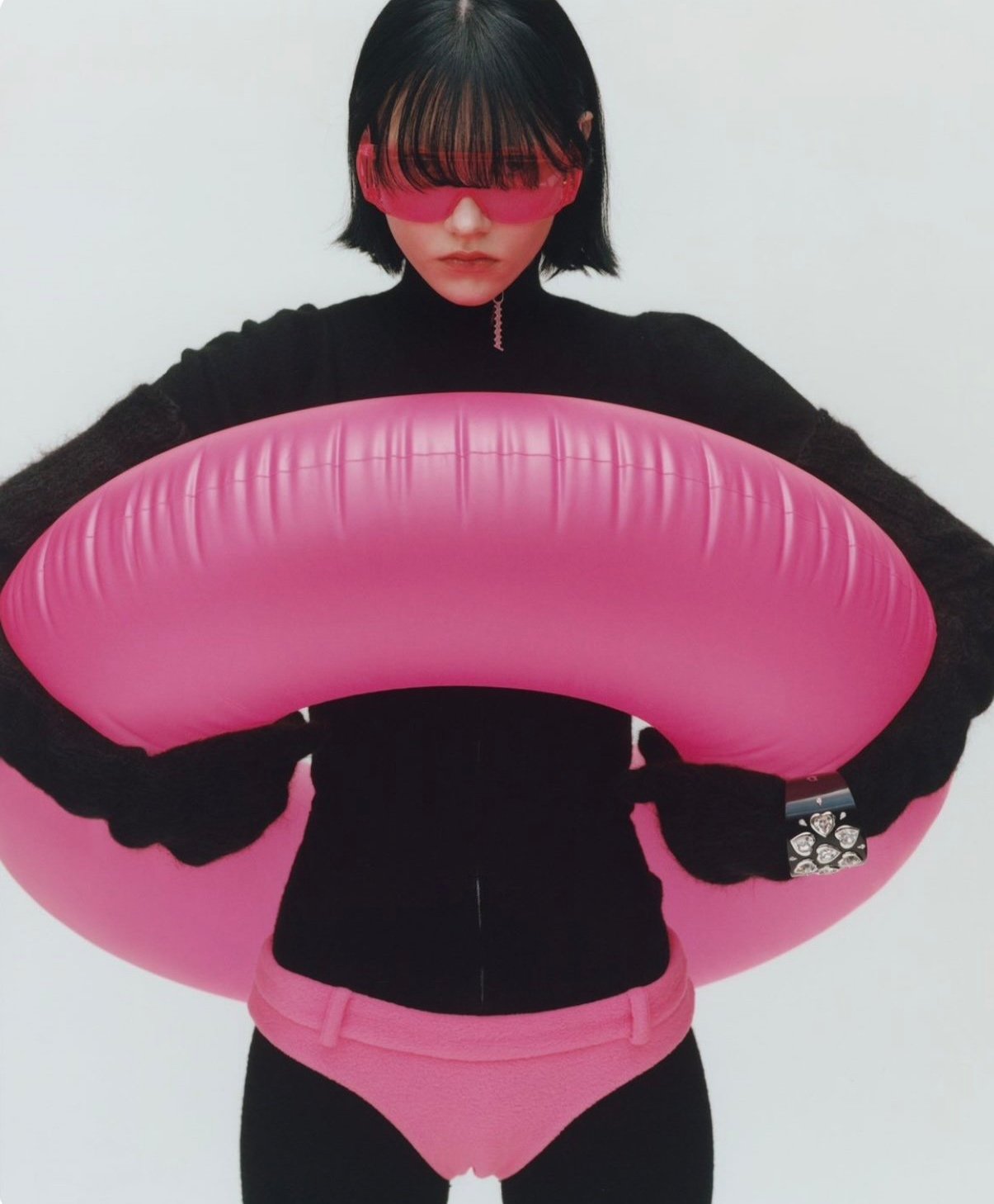 Woman wearing black outfit and pink underwear, holding a large pink inflatable ring with a reflective pink visor on her eyes.