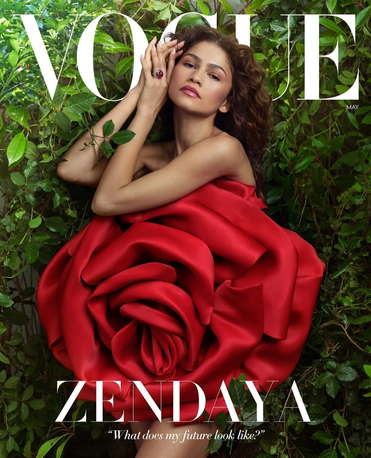 A young woman with curly hair lying among green foliage, dressed in a strapless red fabric that resembles a large rose. She has a ring with a red gemstone on her left hand, which is resting on her forehead. The magazine cover features the text 'VOGUE