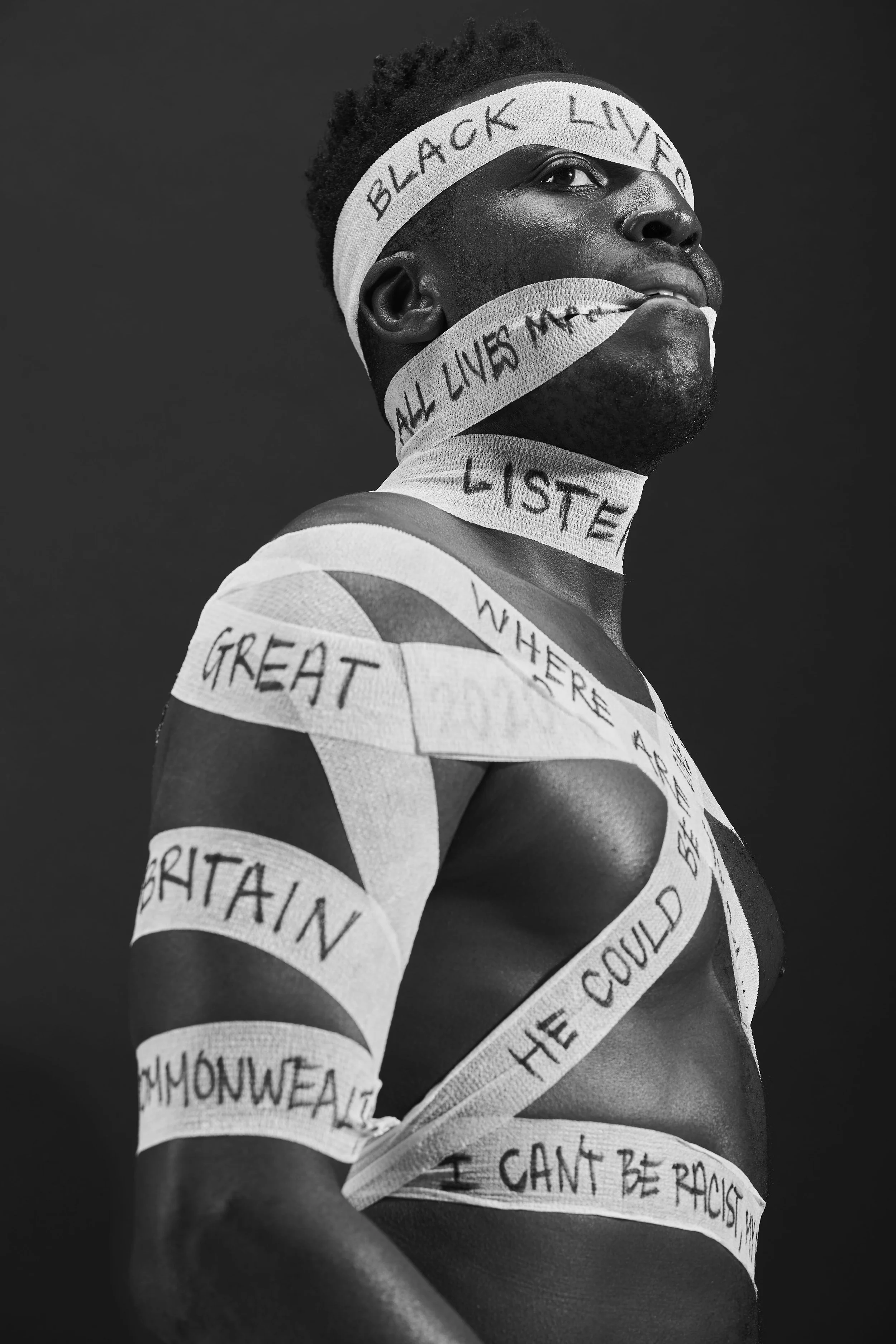 Black and white portrait of a person with their face and body wrapped in strips of white tape with handwritten messages about racial justice and equality, against a plain dark background.