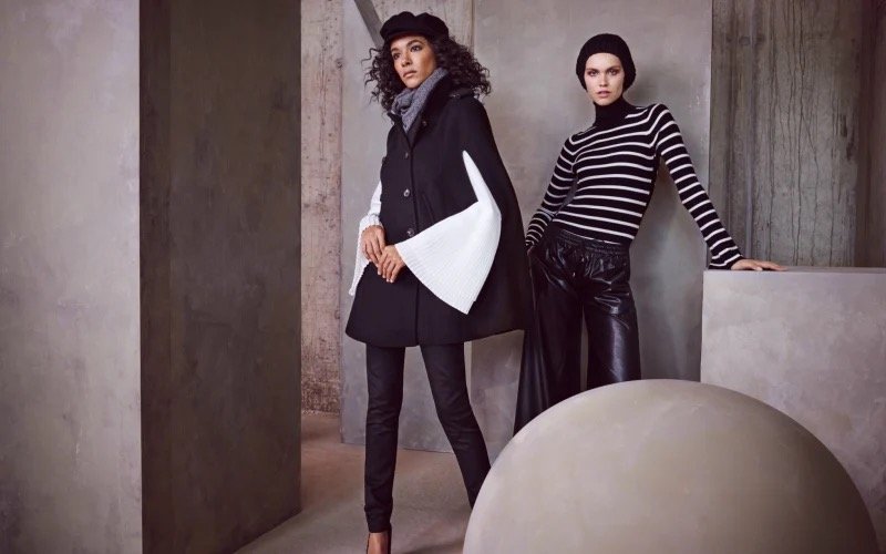 Two women pose in a modern, industrial-style setting with concrete walls. One wears a black skirt and vest with a gray scarf, while the other wears a black and white striped long-sleeve shirt and black leather pants.