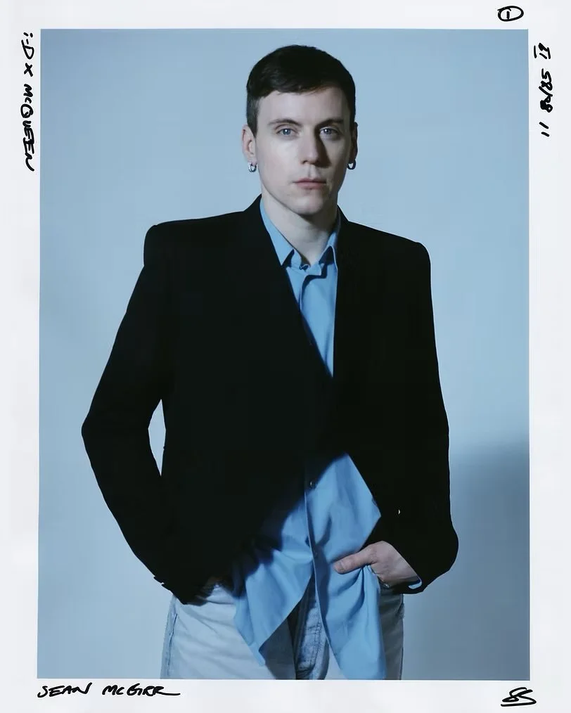 Portrait of a young man with short dark hair, dressed in a black blazer and a light blue shirt, standing against a plain gray background.