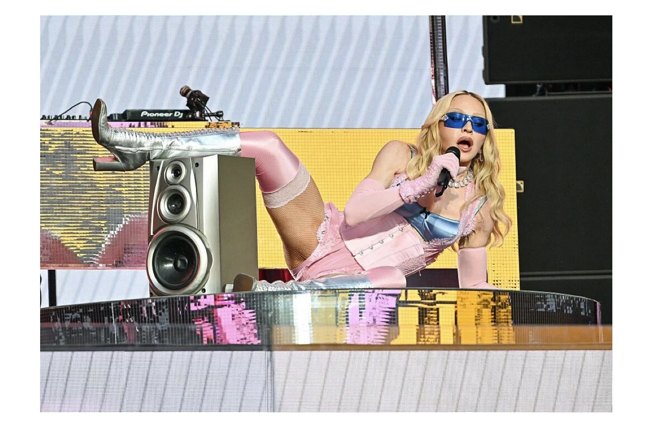 Madonna Time Square performance for Pride. Pink-clad performer lying on a reflective stage with a microphone in hand, wearing blue sunglasses, pink gloves, and metallic silver boots, with DJ equipment and large speakers in the background.