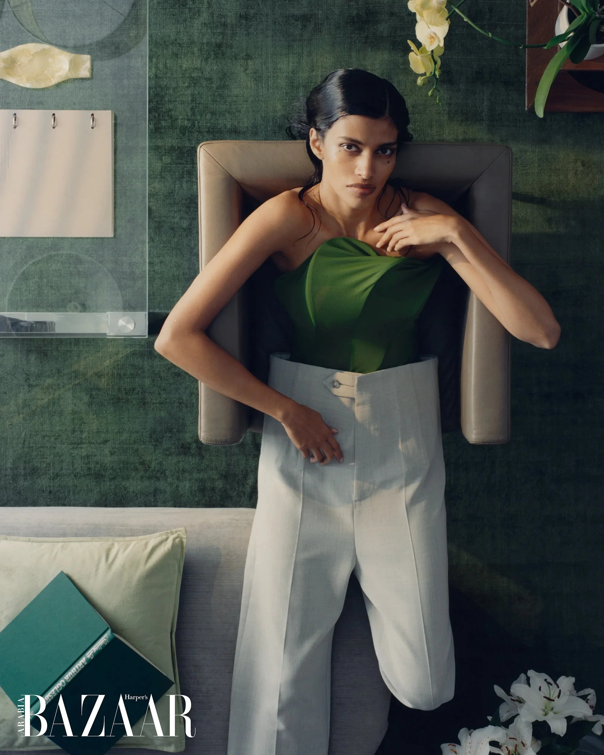 A woman with dark hair styled in a vintage look is sitting in a beige chair, wearing a green top or dress, surrounded by flowers and magazines, with a Harper's Bazaar magazine visible in the corner.