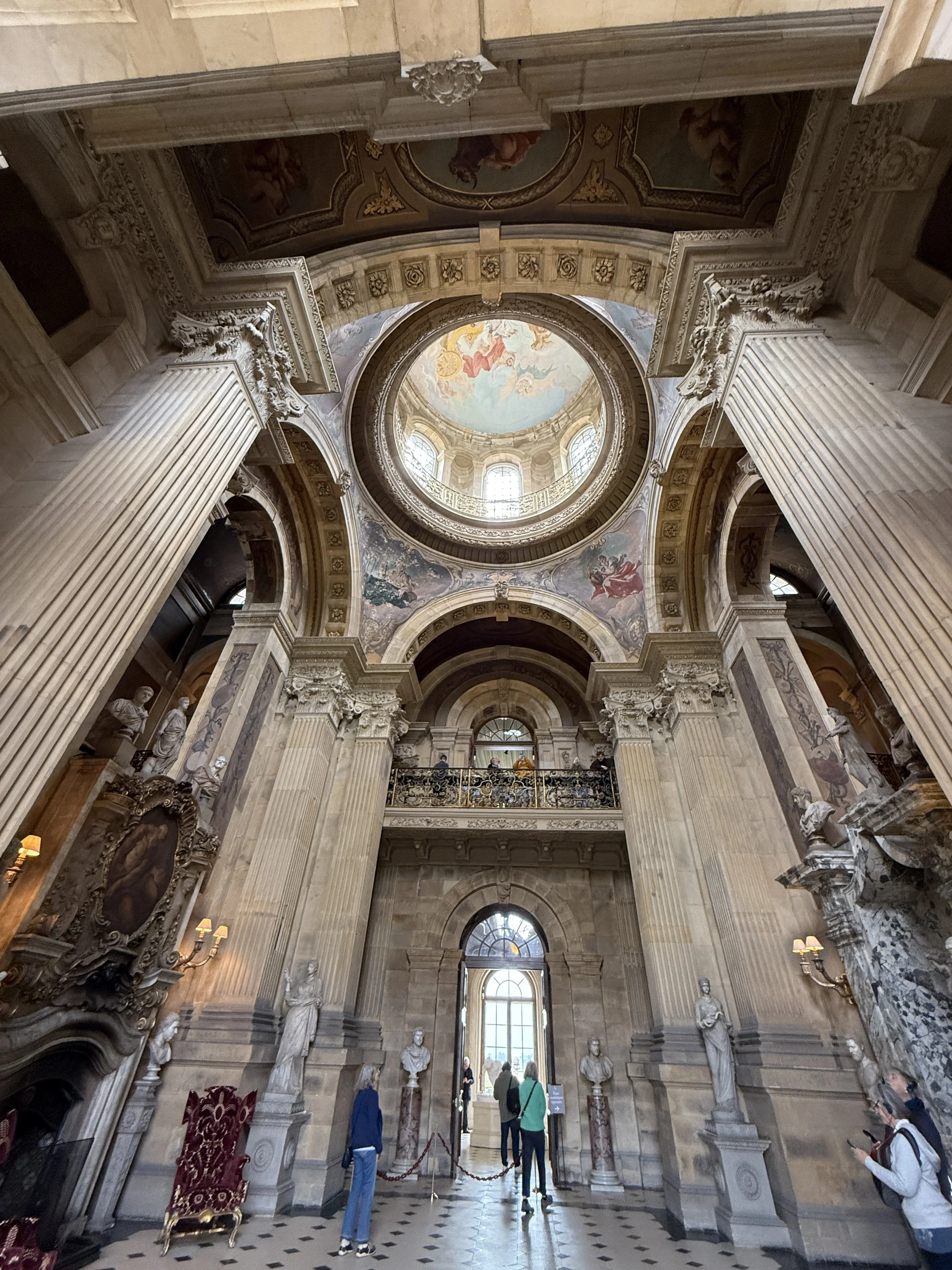 View of the interior of a grand historic building with high ceilings, ornate columns, statues, and frescoes, with several people sightseeing.