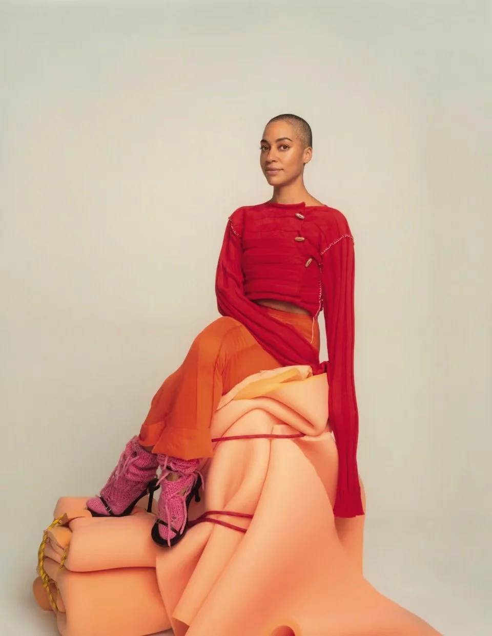 A woman with a shaved head sitting on a surreal sculpture of a human body, wearing a red long-sleeved top, orange skirt, pink knitted boots, and black shoes.