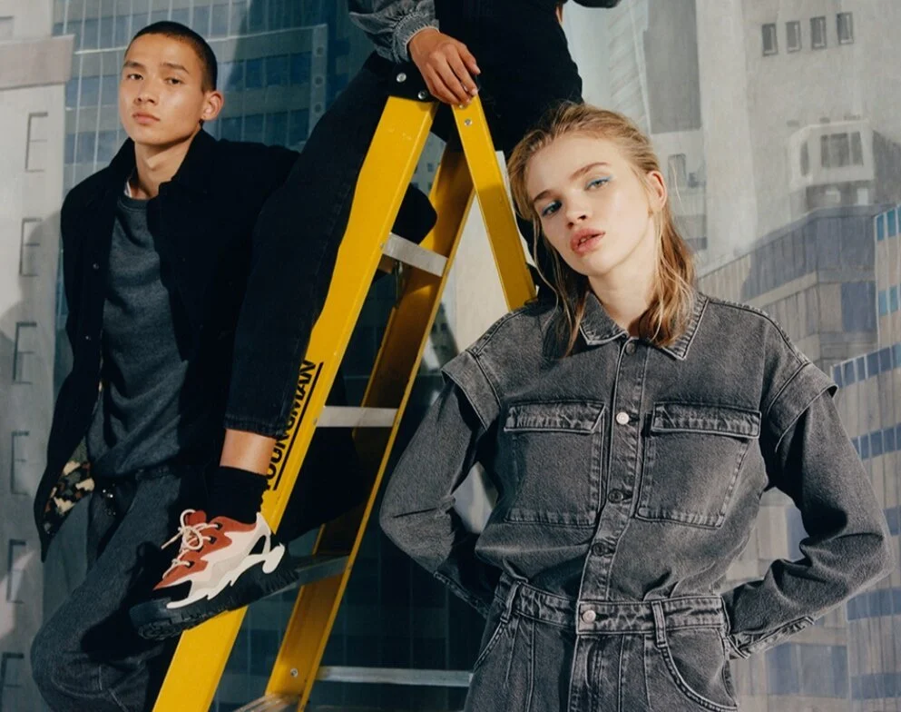 A young man and woman posing in front of a city skyline backdrop. The man is sitting on a yellow ladder, and the woman stands with her hands on her hips.
