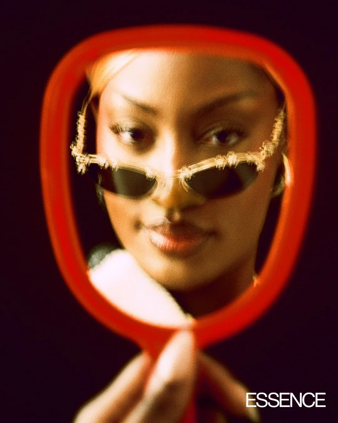 A woman looking into a handheld mirror with rounded edges, wearing sunglasses and posing against a dark background, with the word 'ESSENCE' in the bottom right corner.