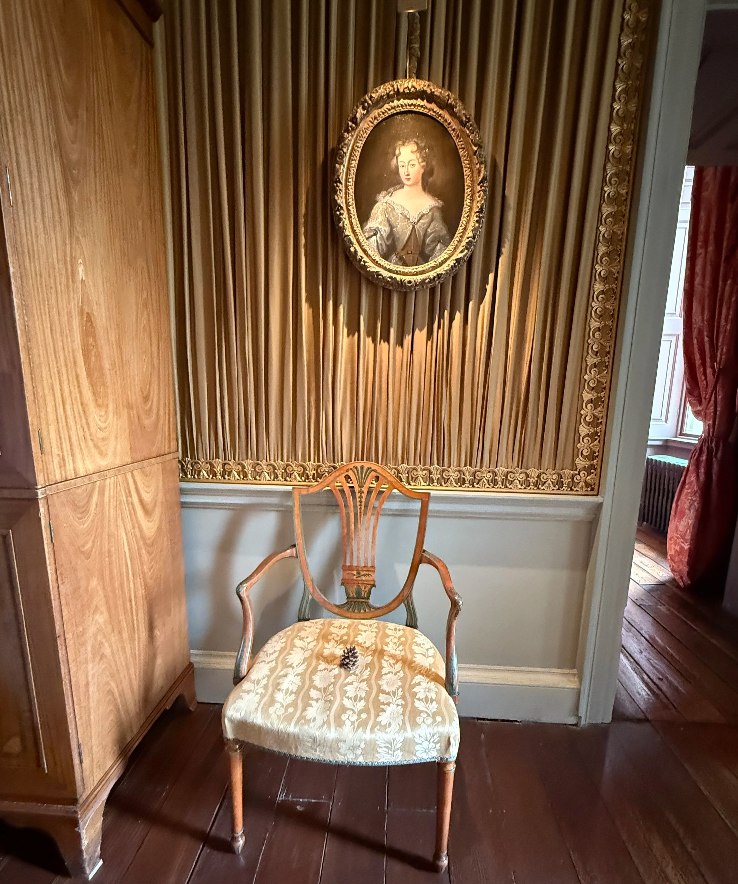 An antique wooden chair with a floral upholstered seat and ornate carved backrest, placed in a room with wood-paneled walls and a framed portrait of a woman hanging above it.
