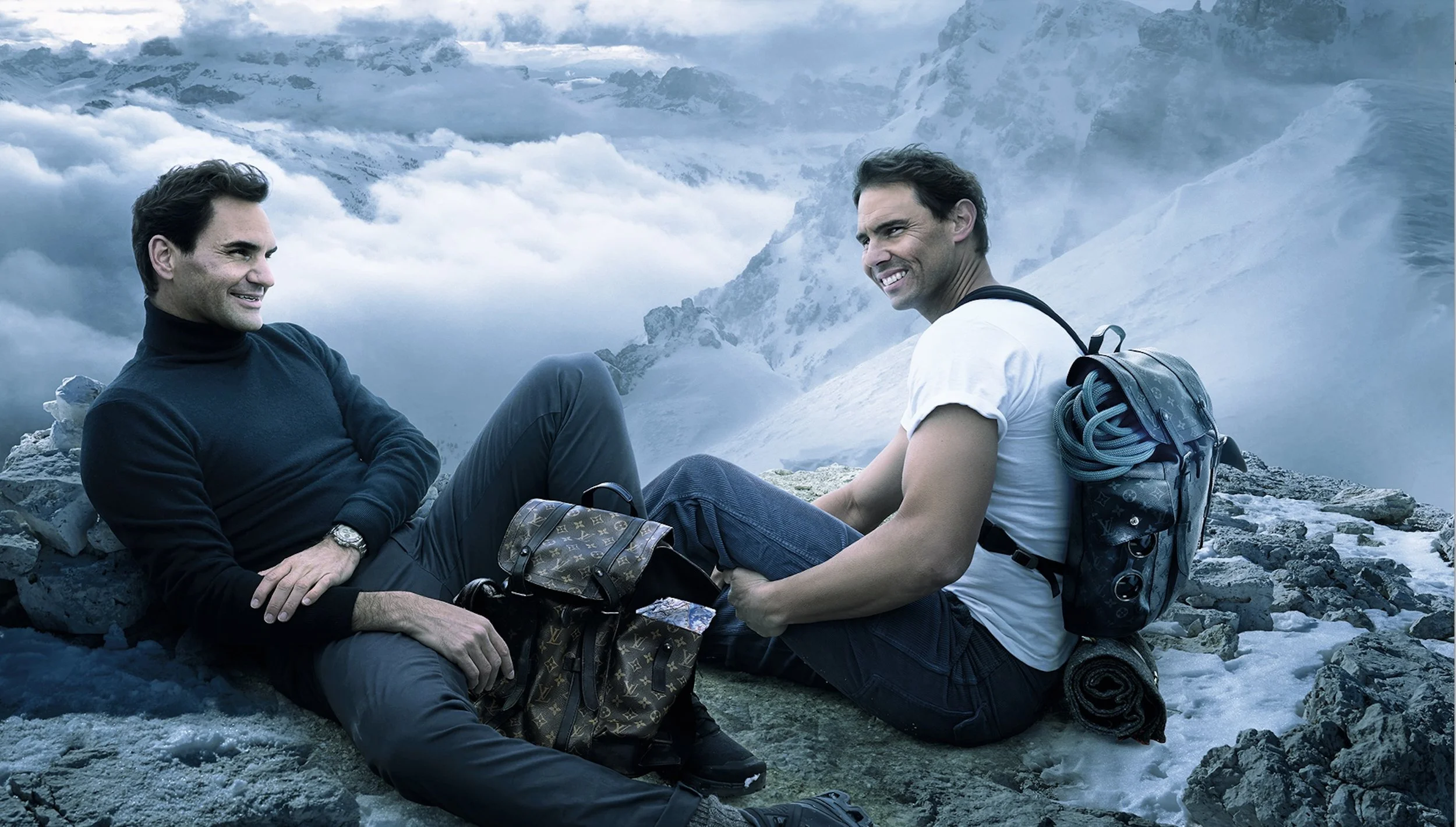Two men relaxing on rocky mountain terrain with snow-covered peaks in the background, smiling and enjoying the scenic view.