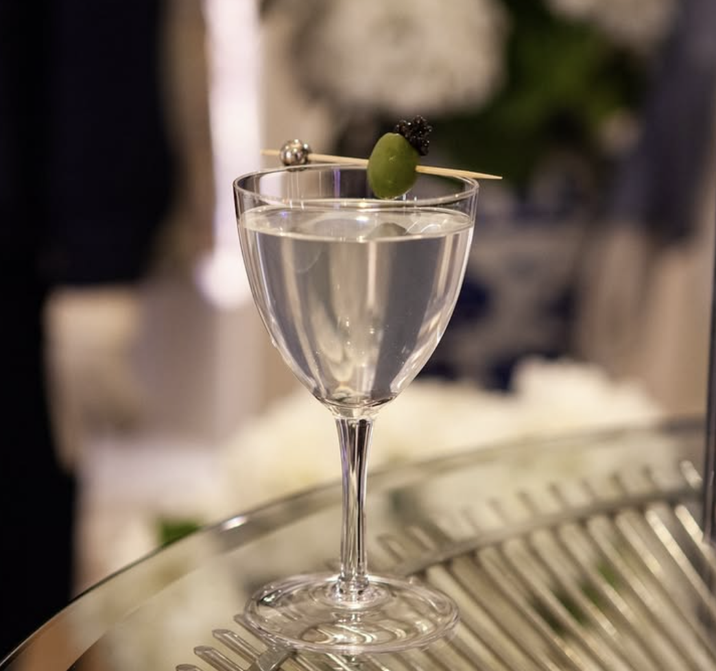 A cocktail glass filled with a clear drink, garnished with an olive and a black berry on a skewer.