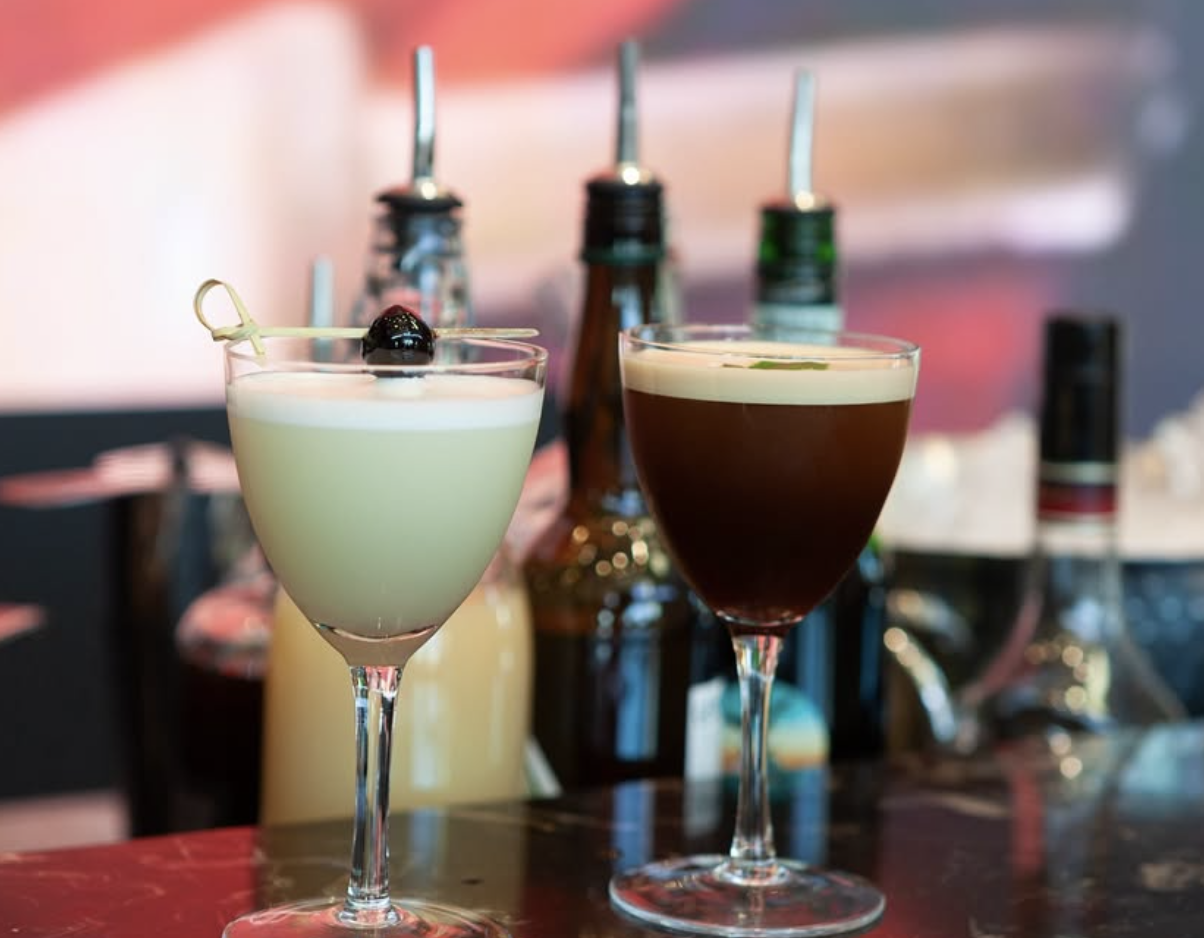 Two cocktails on a bar, one with a cherry garnish, behind bottles of syrups and alcohol.