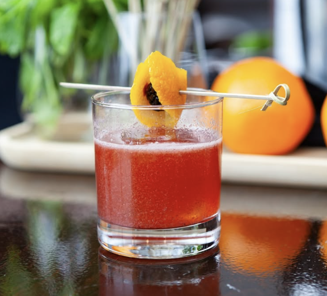 A glass of red cocktail garnished with a slice of orange and a blackberry on a skewer, with oranges and a blurred background behind it.