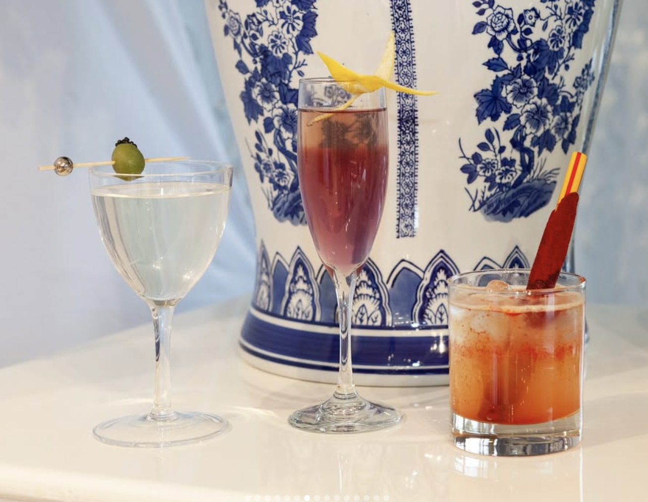Three cocktails on a white surface with a blue and white decorative background. The left drink is a clear cocktail in a wine glass garnished with a green olive. The middle drink is a pinkish cocktail in a champagne flute with a lemon twist garnish. T