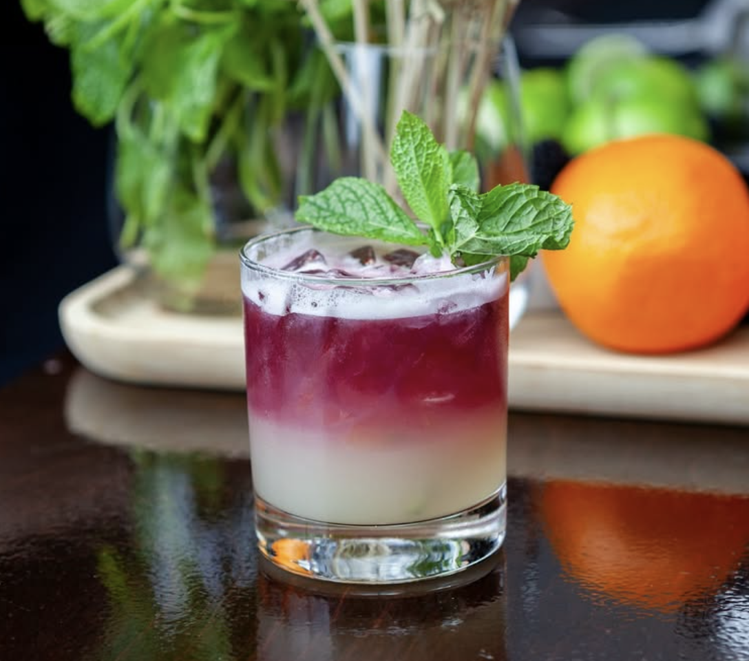 A layered cocktail in a glass with a gradient from white at the bottom to purple at the top, garnished with fresh mint leaves.