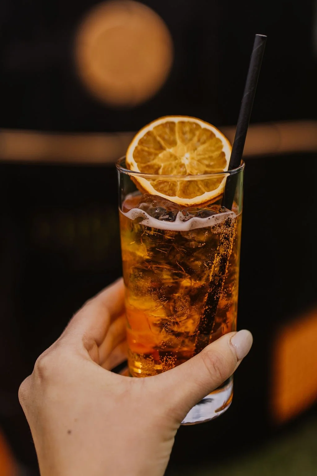 A hand holding a glass of iced orange soda garnished with a slice of dried orange, with a black straw in the drink.
