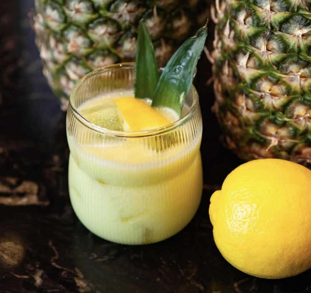 A glass of pineapple and lemon juice with pineapple slices and whole pineapples in the background, and a lemon placed beside the glass on a dark surface.