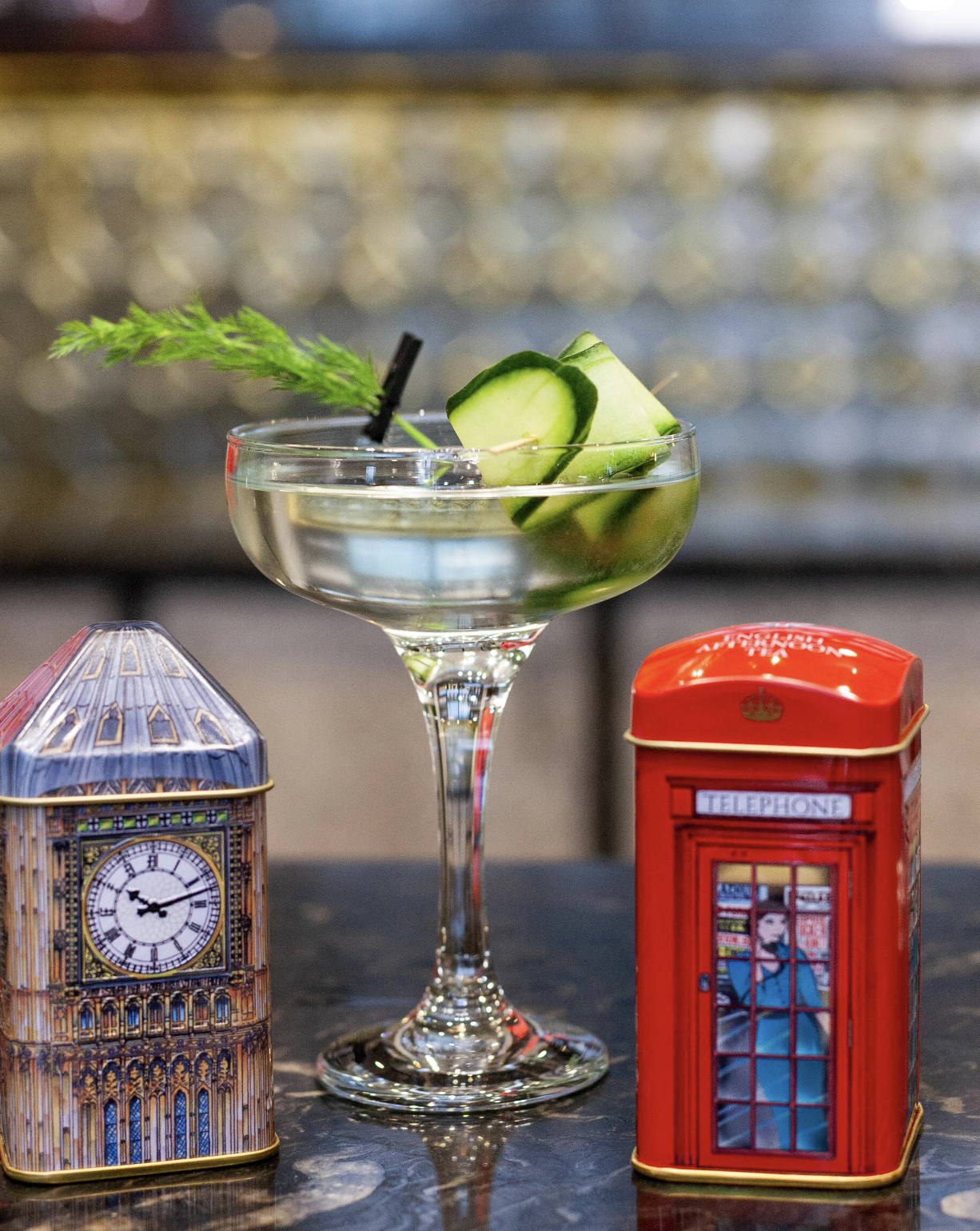 A cocktail glass with cucumber slices and a green garnish, flanked by two decorative tins designed as London's Big Ben and a red telephone booth.