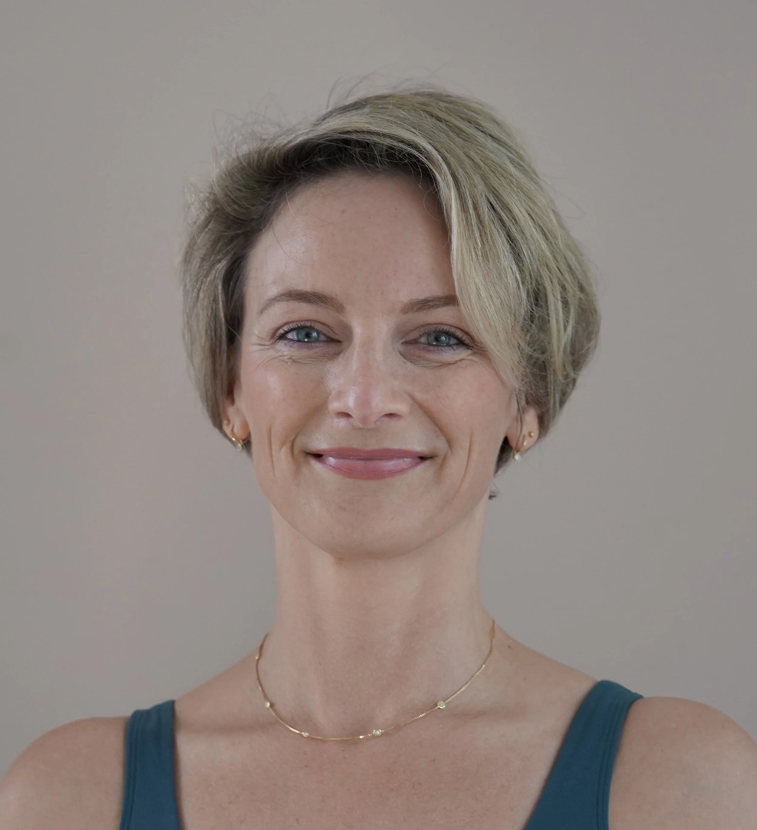 Close-up portrait of a smiling woman with short blonde hair, blue eyes, wearing a blue top, small earrings, and a delicate gold necklace, standing against a plain background.