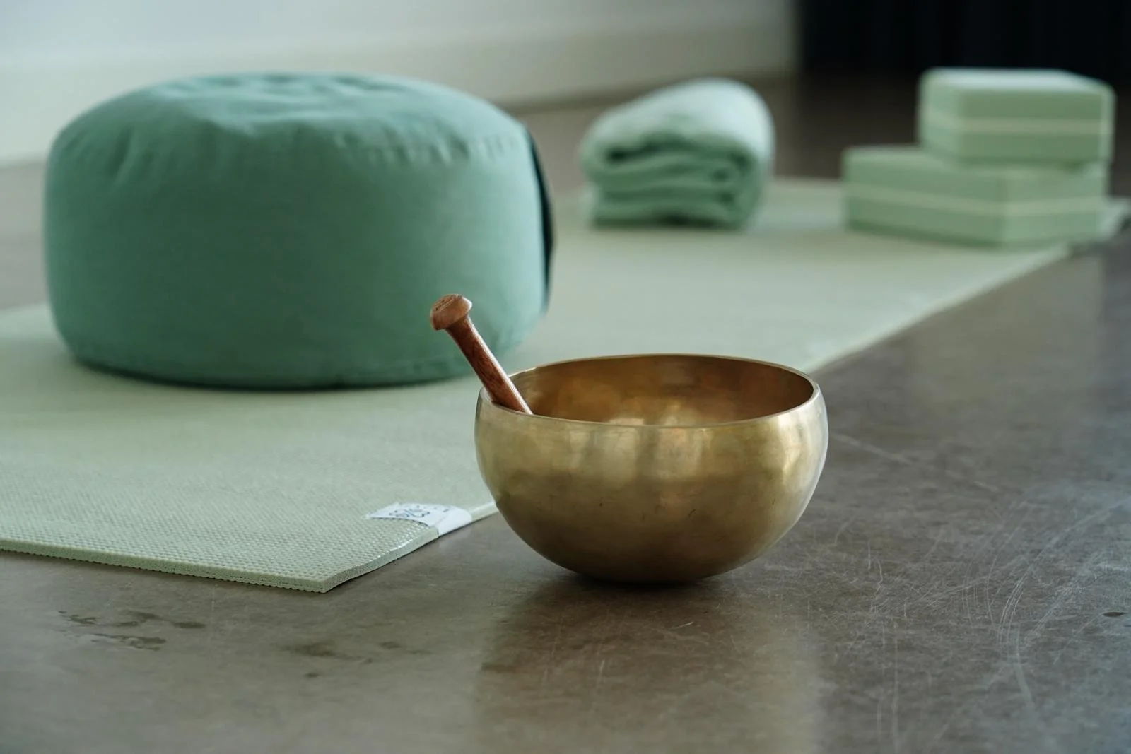 A brass singing bowl with a wooden mallet inside, placed on a light green meditation mat on a wooden surface, with folded green linens and a green cushion in the background.