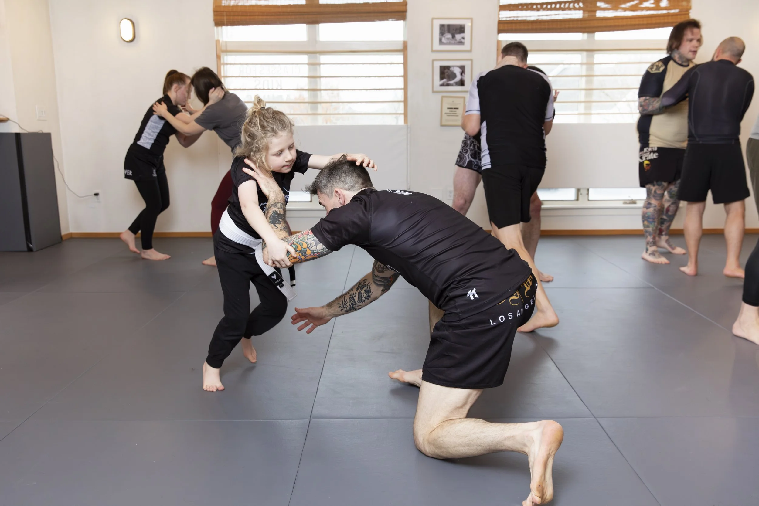 Martial arts class with children and adults practicing grappling techniques on a padded floor in a bright room.