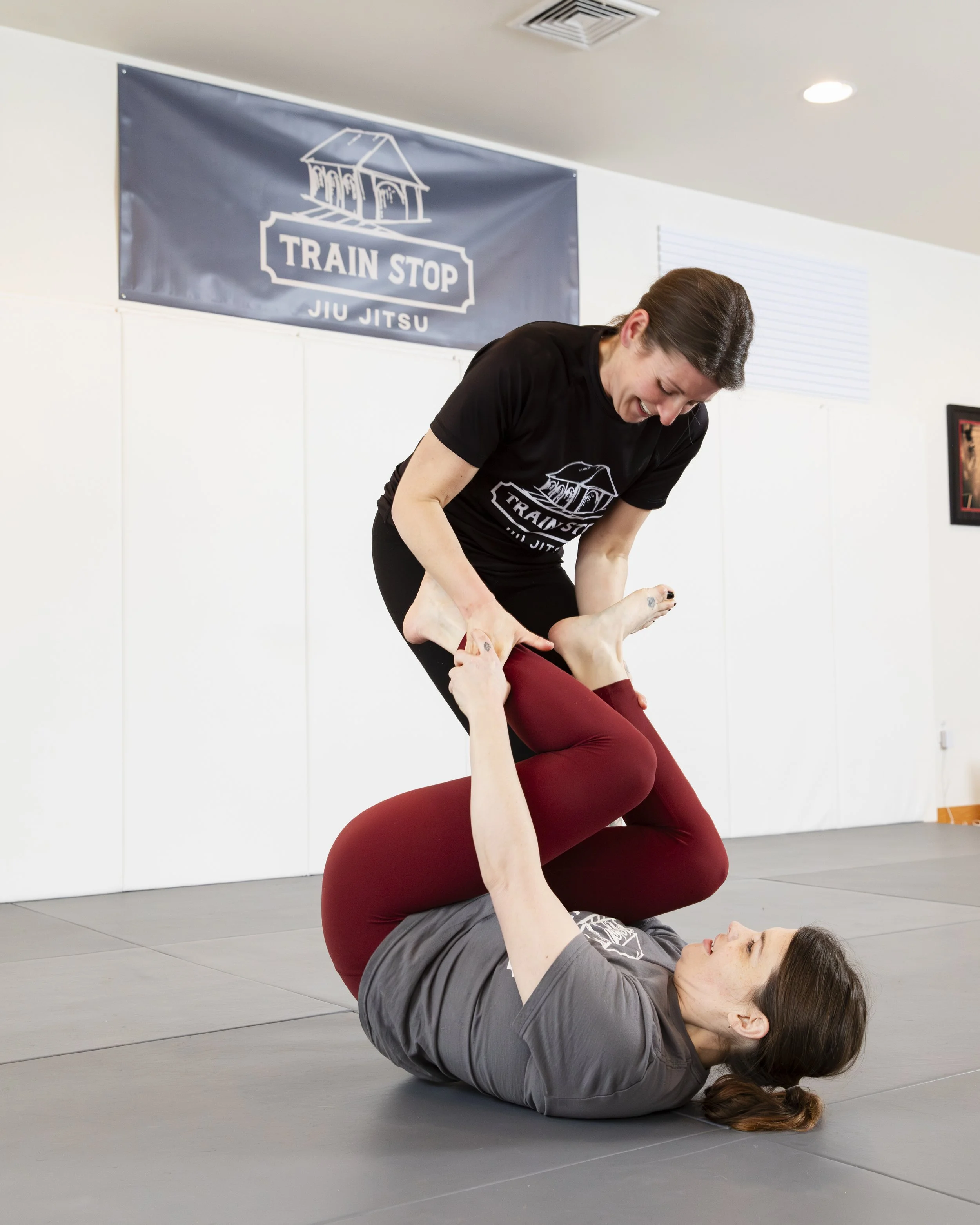 Two women practicing Brazilian Jiu-Jitsu in a training gym, with one woman on the ground holding the other in a submission move, and a "Train Stop Jiu Jitsu" banner on the wall.