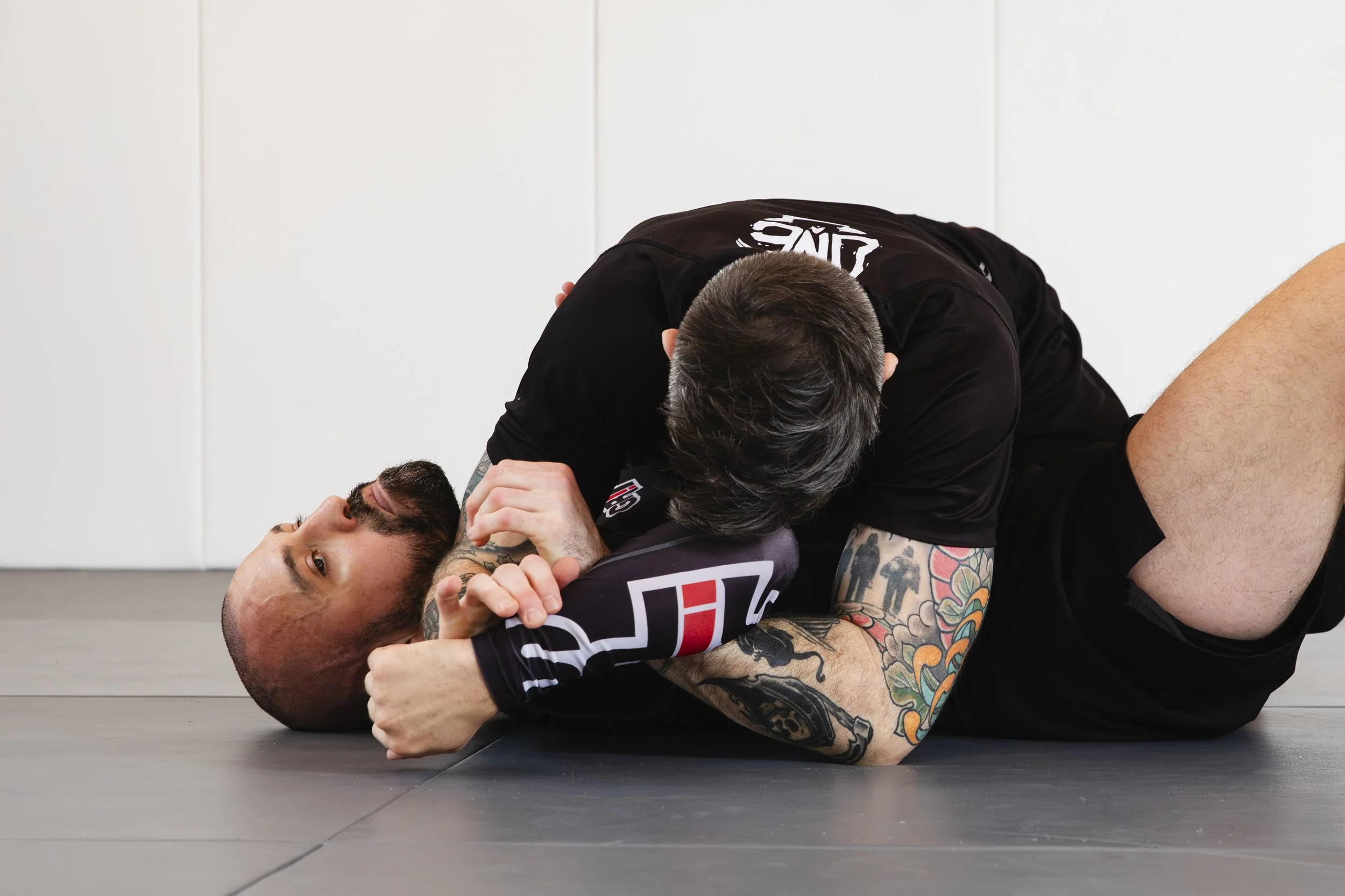 Two men practicing Brazilian Jiu-Jitsu, with one man on top in a dominant position on the mat while the other man is on the ground with his head turned and looking at the camera.