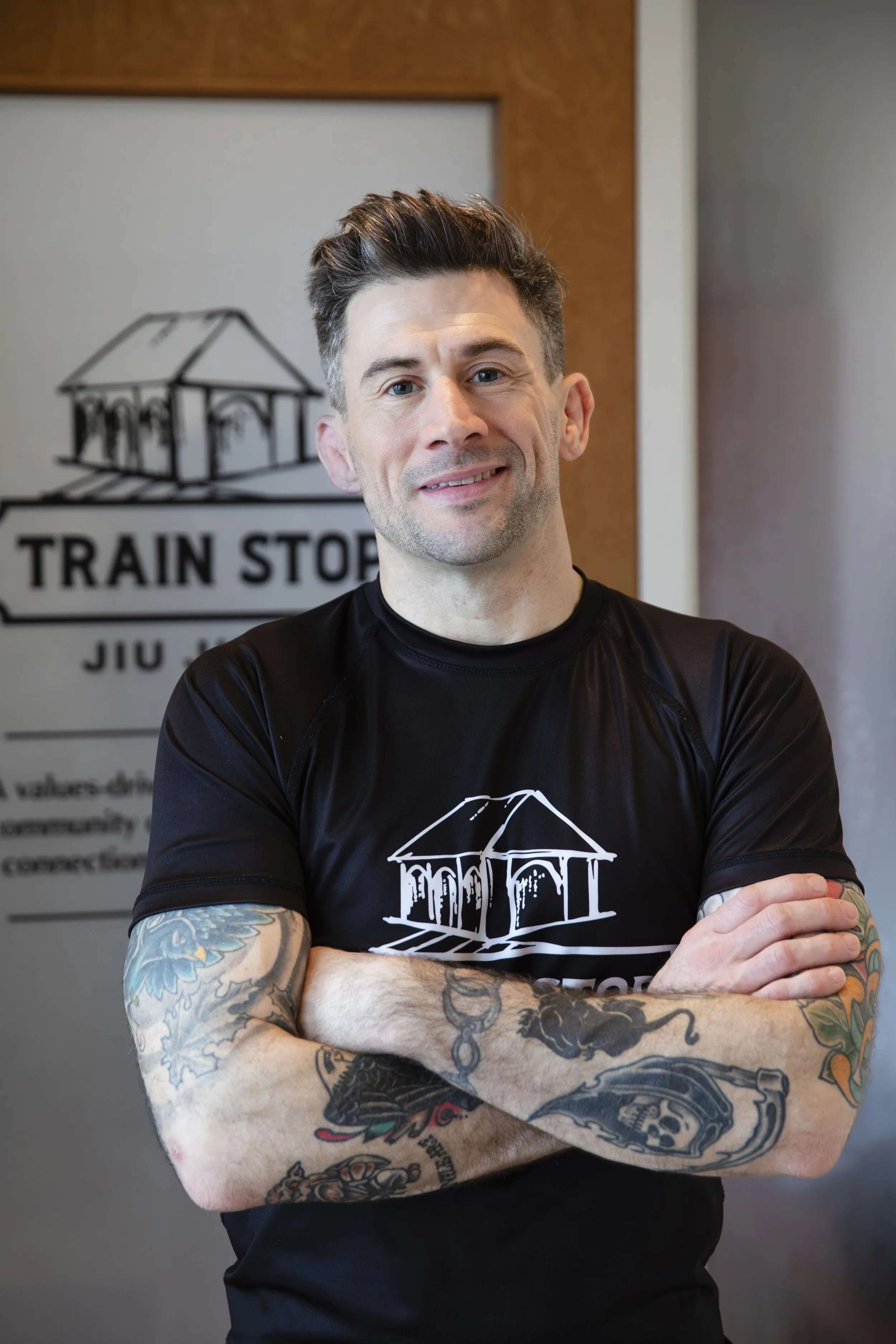 A tattooed man in a black T-shirt stands with arms crossed in front of a logo that reads 'Train Stop Jiujitsu' on a wall.