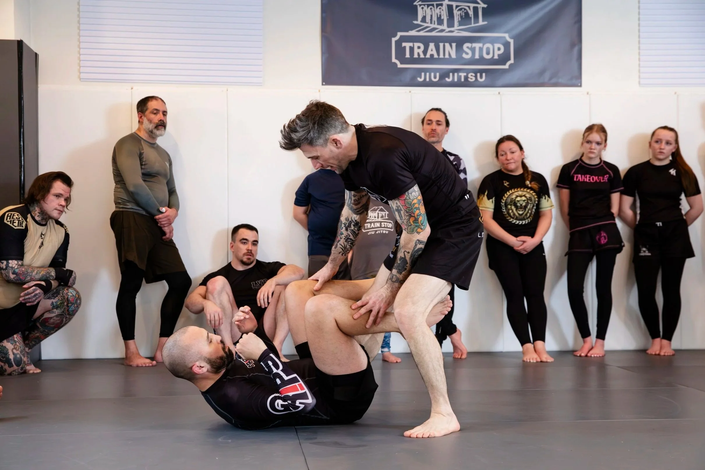 Two men practicing Brazilian Jiu Jitsu on a mat, with one man on the ground in a black rash guard and shorts, and the other man standing over him, gripping his legs. Several people watch in the background inside a gym with a banner that reads 'Train Stop Jiu Jitsu.'