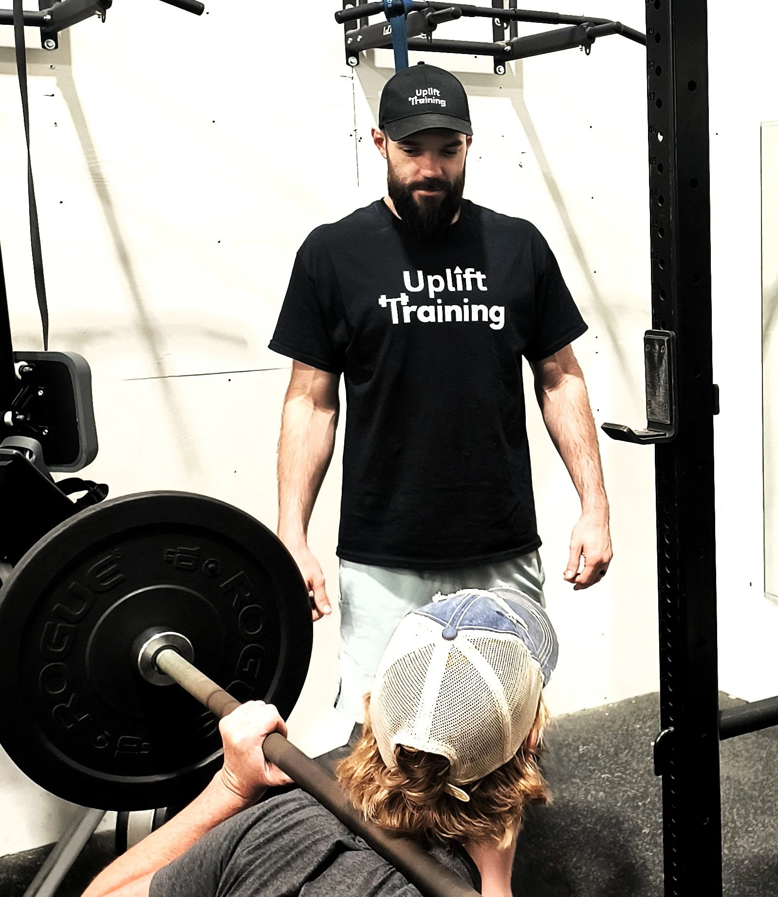 A man in a black 'Uplift Training' T-shirt and cap observes a woman in a gray shirt and baseball cap performing a barbell bench press in a gym.