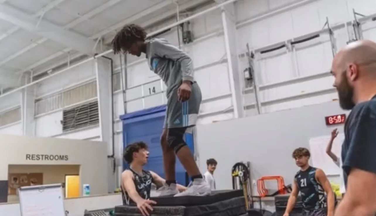 A young man is balancing on a stack of tumbling mats during a gym training session, with a coach and other athletes nearby.