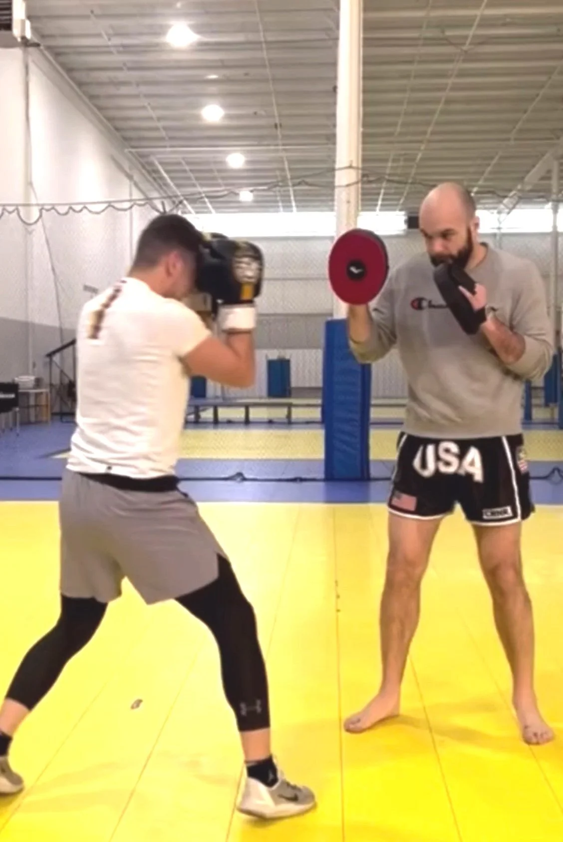 A man in boxing shorts, labeled USA, is training with a pad held by a trainer, while another person is sparring with boxing gloves in an indoor gym with yellow mats.