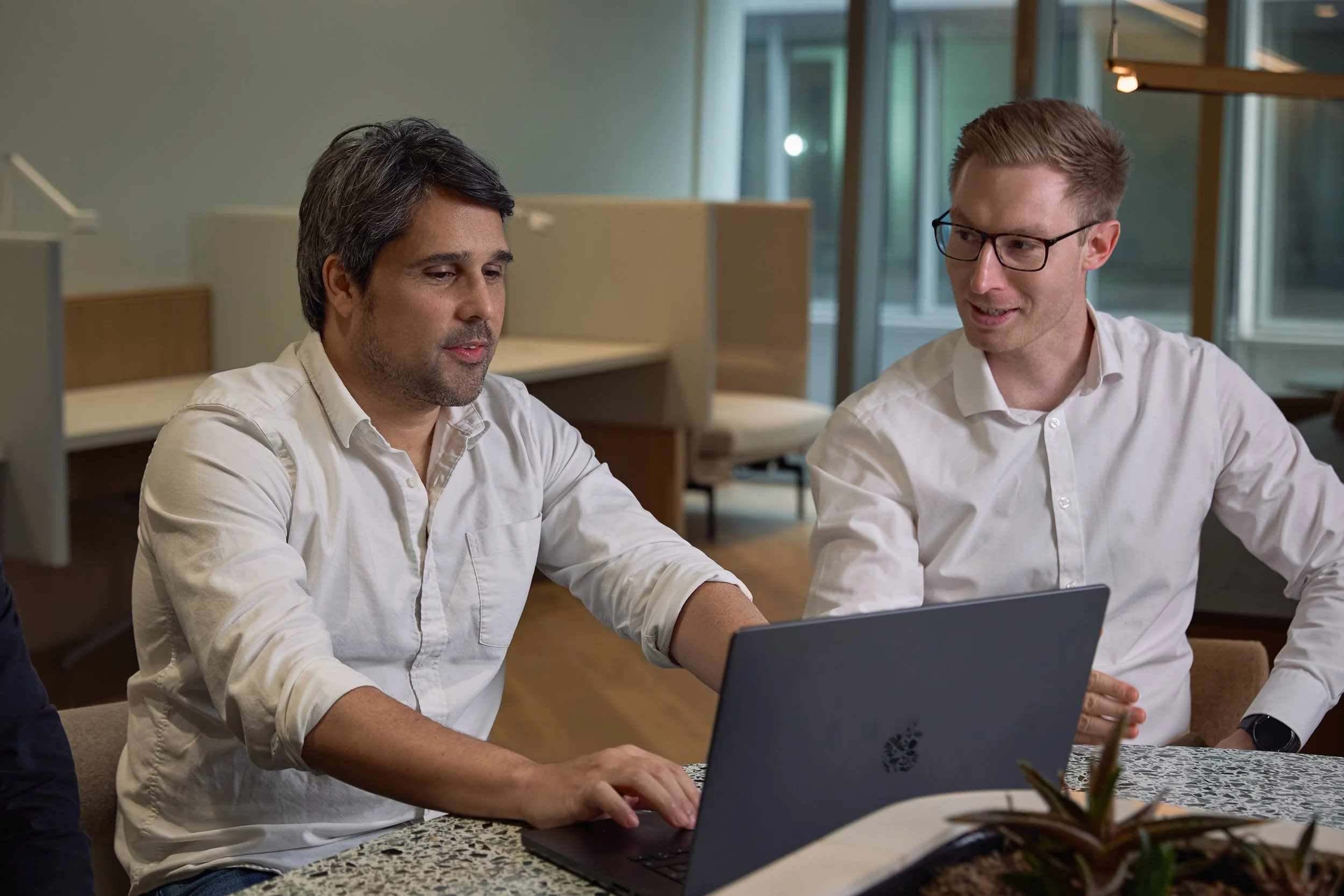 Two men in white shirts sitting at a table with a laptop, engaged in discussion in a well-lit modern office.