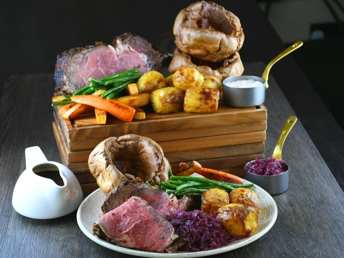 A traditional Sunday roast with sliced beef, roasted potatoes, carrots, green beans, and mushrooms, served on a white plate. The beef, potatoes, carrots, and green beans are displayed on a wooden serving board with sauces in small metal containers, on a dark wooden table.