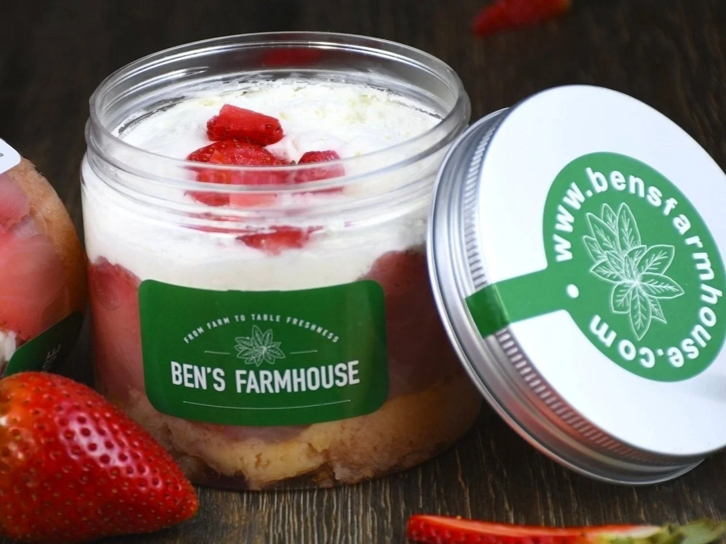 A clear plastic container of strawberry shortcake with a green label that reads 'Ben's Farmhouse', topped with strawberries and whipped cream, sitting on a dark wooden surface with a strawberry and a sliced strawberry beside it.