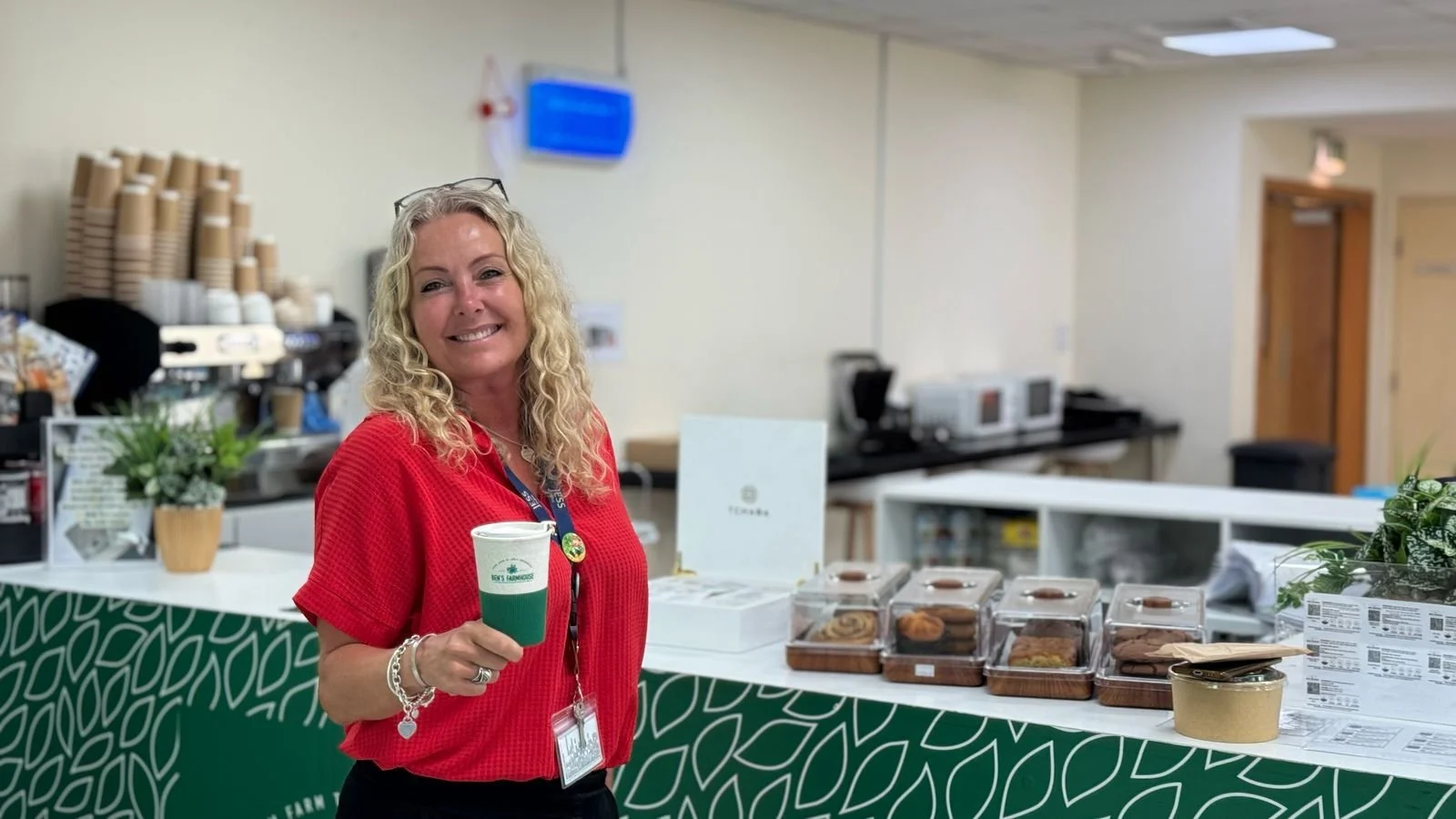 A woman with curly blonde hair, wearing a red shirt, smiling and holding a disposable coffee cup, standing in front of a table with baked goods and promotional materials.