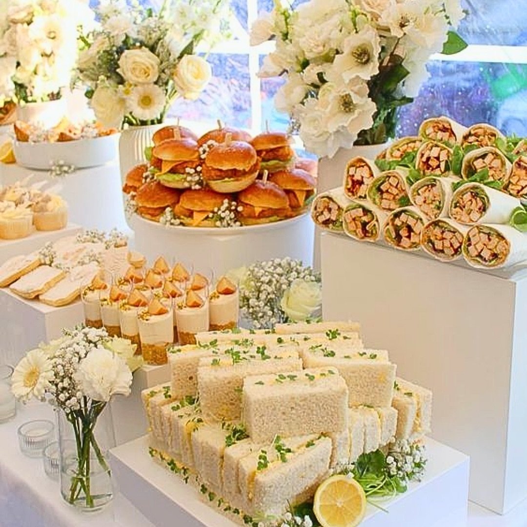 A celebration table with sandwiches, mini cakes, and wraps, decorated with white flowers and lemon slices.