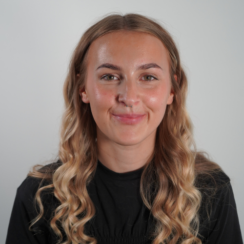 A woman with long, wavy blonde hair and a nose ring, smiling at the camera against a plain light gray background.