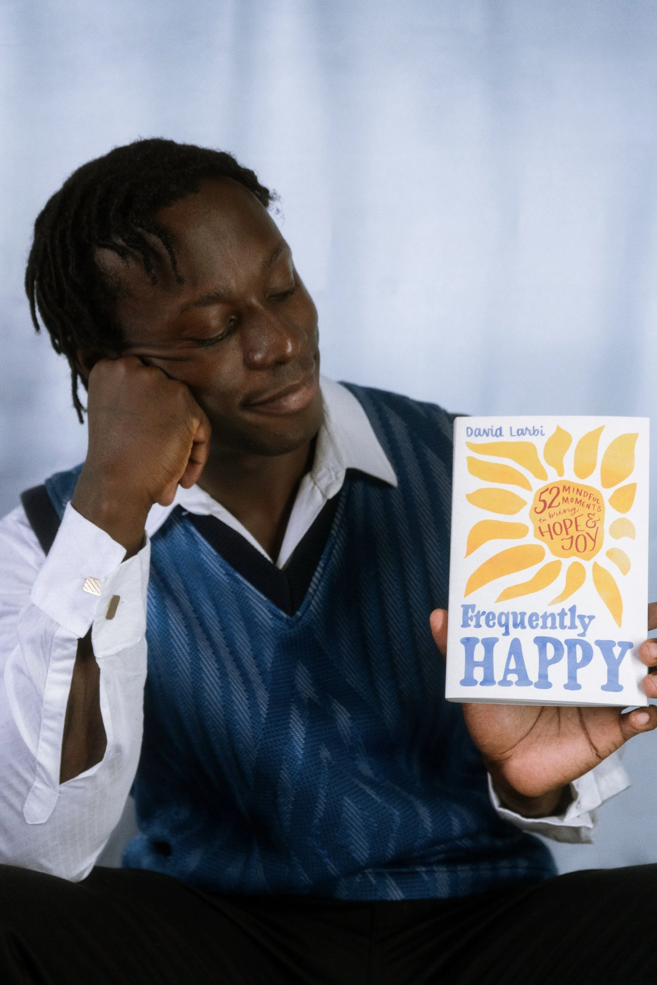 A man with dark skin and dreadlocks sits with his head resting on his hand, wearing a white shirt and a blue vest, holding a colorful card with the word 'happy' visible.
