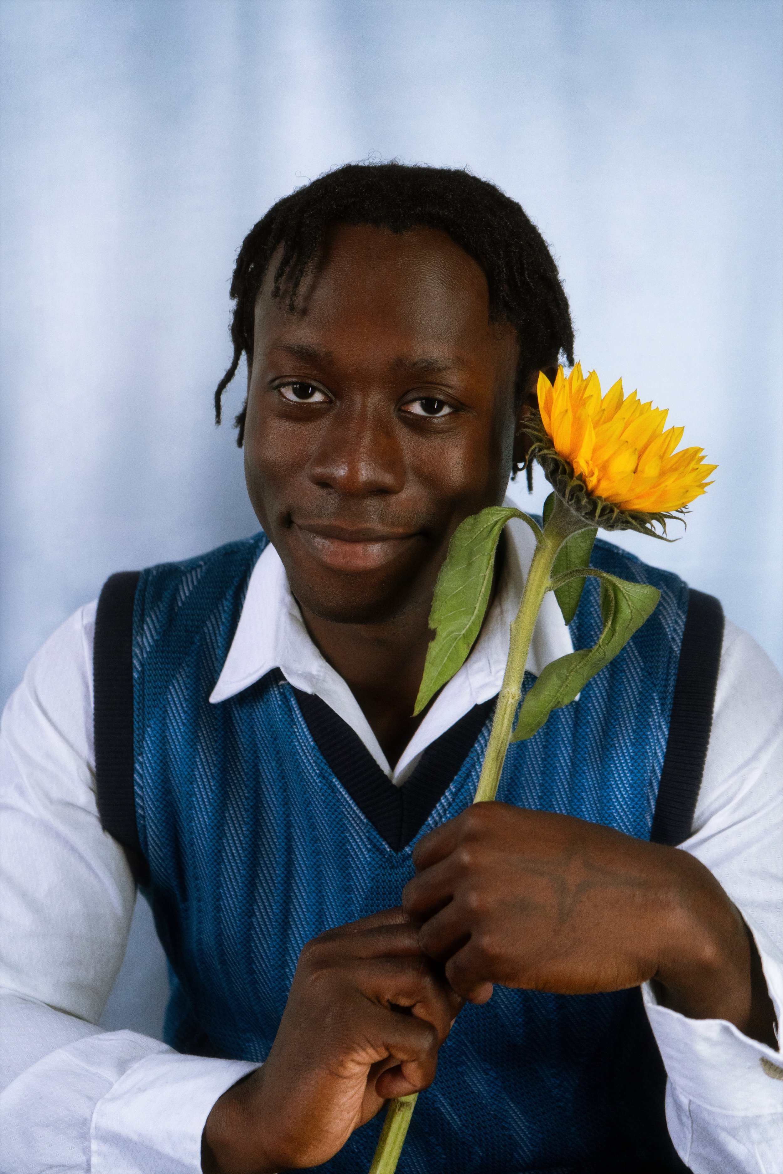 A young man in a white shirt and blue sweater vest holding a sunflower, sitting against a light blue background.