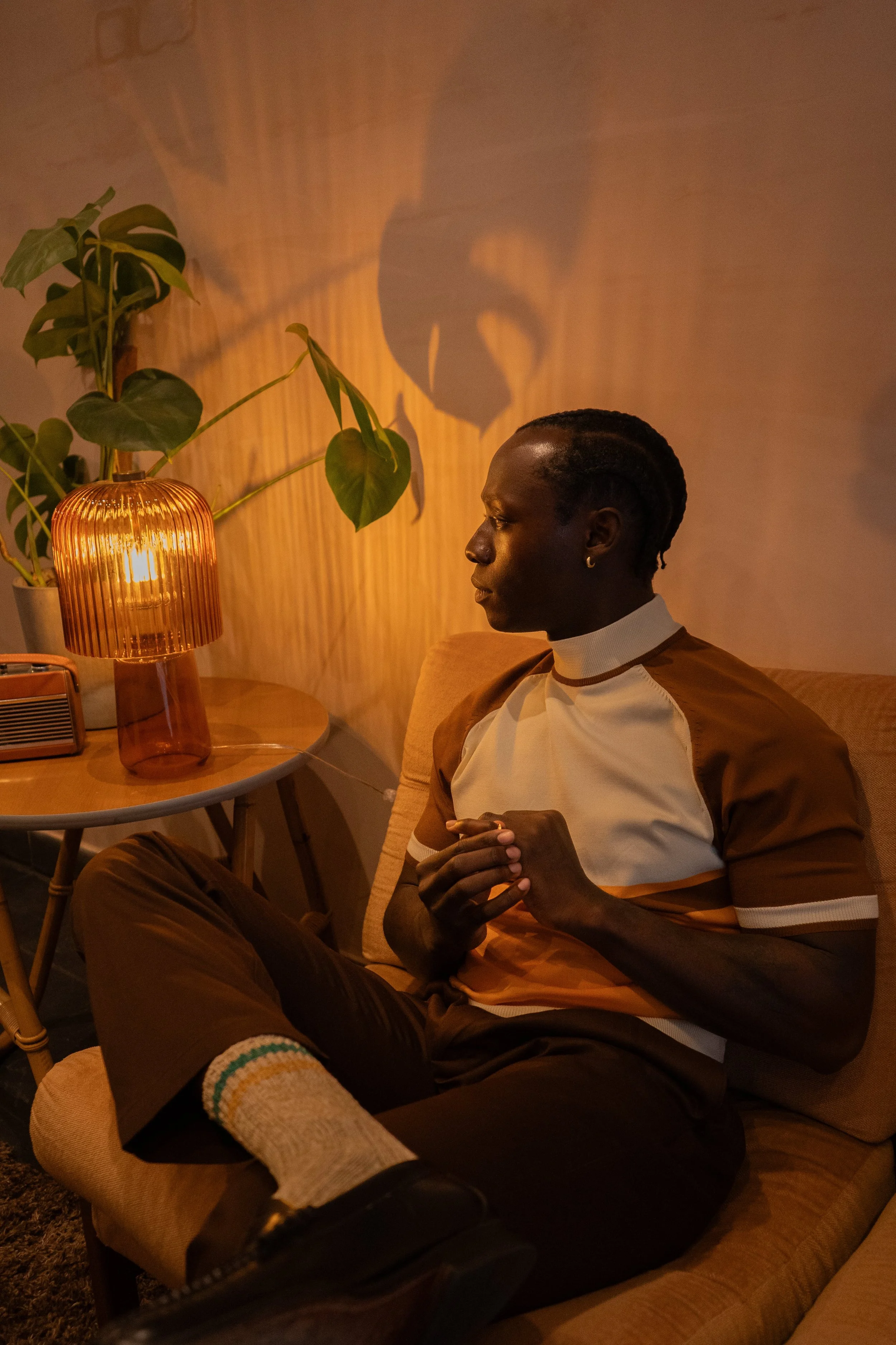 A man sitting on a beige couch with his legs crossed, clasping his hands. He is wearing a short-sleeved shirt with brown and white colors, brown pants, and beige socks with rainbow stripes. Next to him, on a round side table, is a orange glass lamp casting a warm glow, and there is a potted plant with large green leaves behind the lamp. The man is looking to the right, with a contemplative expression, in a softly lit room.
