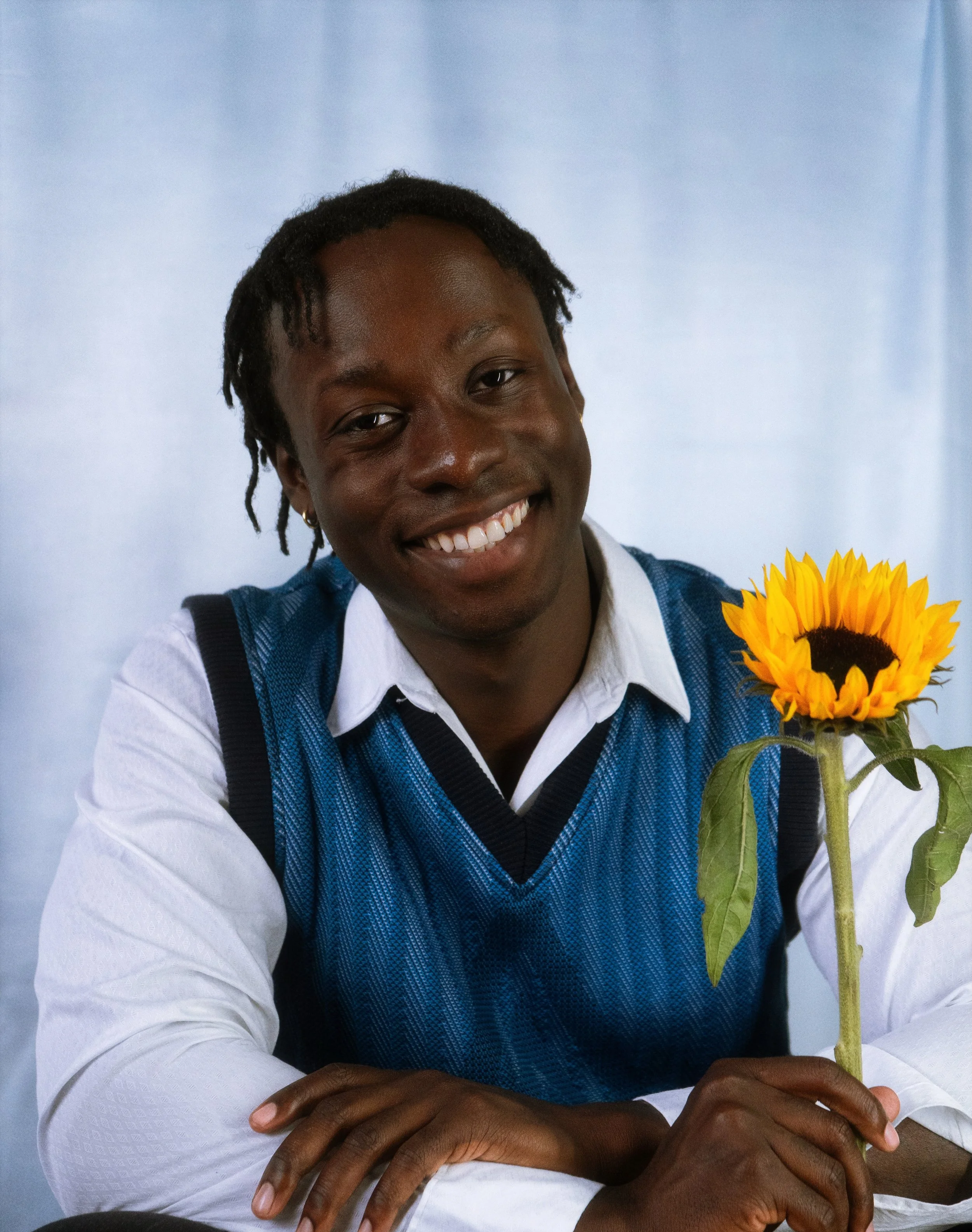 David Larbi wearing a blue vest over a white shirt, holding a sunflower, sitting against a light blue background.