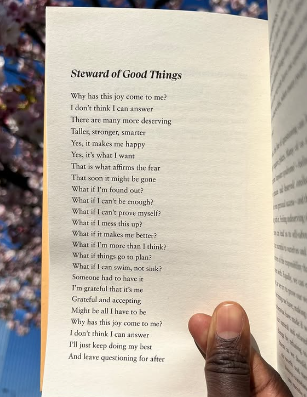 A person holding an open book with a poem titled "Steward of Good Things" on a cream-colored page. The background is blurred with colorful items, possibly in a store or library.