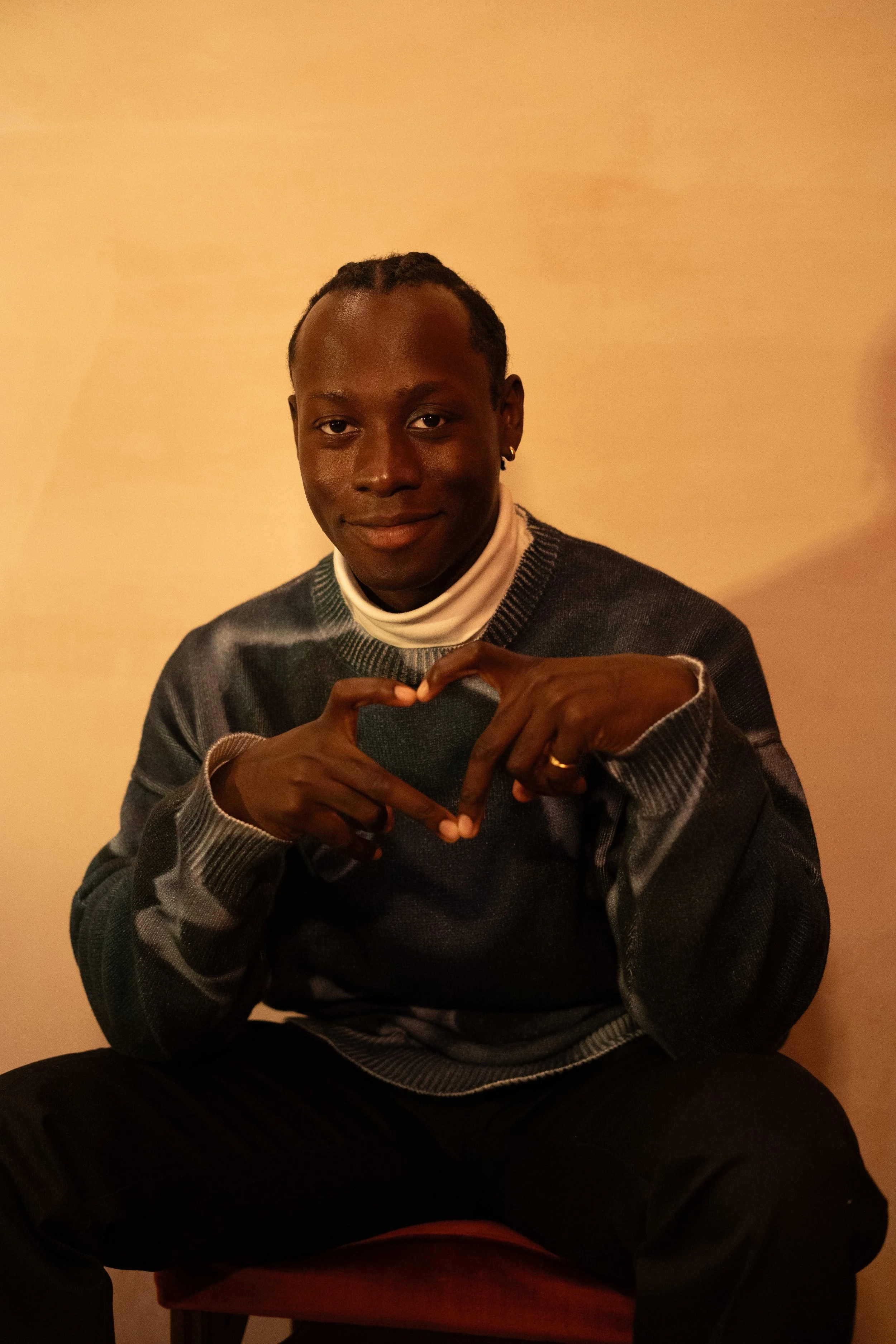 David Larbi sitting on a chair, making a heart shape with his fingers in front of a beige wall.