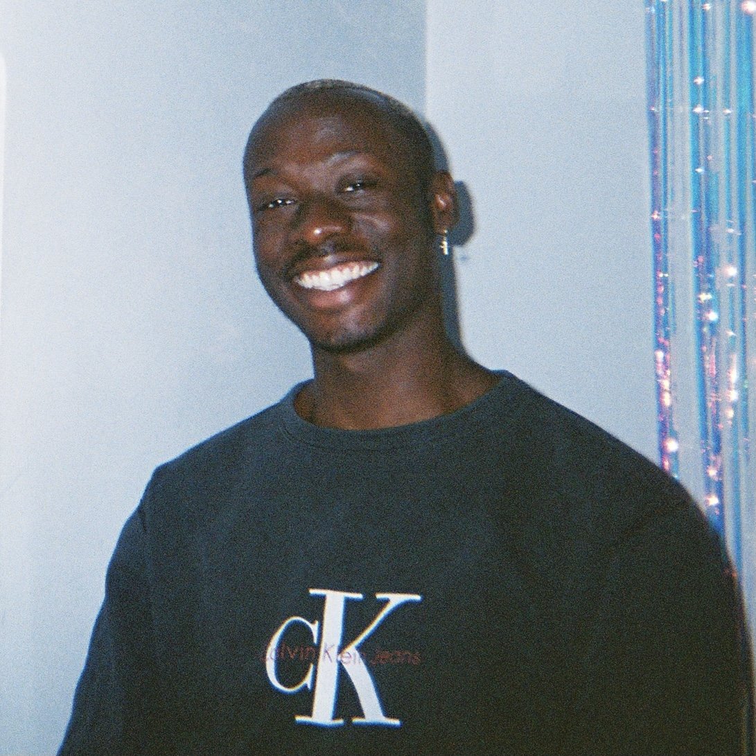 David Larbi wearing a black Calvin Klein t-shirt, standing indoors next to a wall decorated with shiny, multi-coloured streamers.