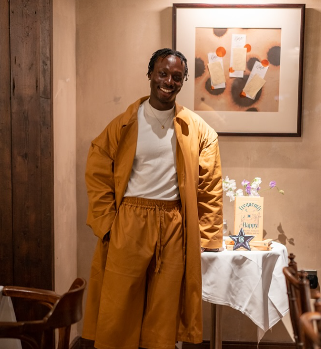 David Larbi in a mustard-coloured coat and matching pants standing indoors near a table decorated with flowers and a cake, with a framed poster of notes pinned to the wall behind him.